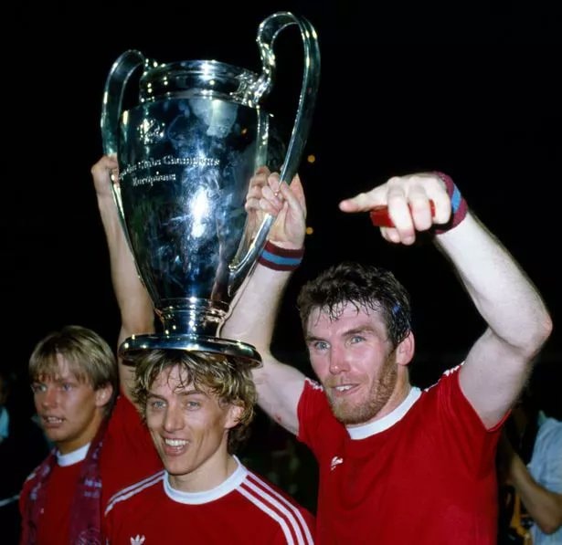 Group of football players celebrating a championship victory while holding the trophy. They are wearing red jerseys and are smiling happily.